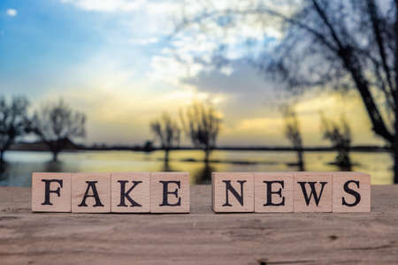 Fake News text written on wooden cube block on wooden table with blurred background of landscape and...