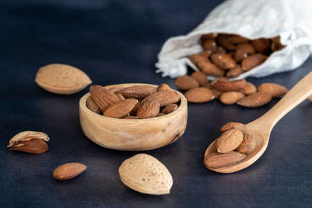 Almonds in a wooden bowl and on a wooden spoon on a black wooden table. some fruits are in a cotton...