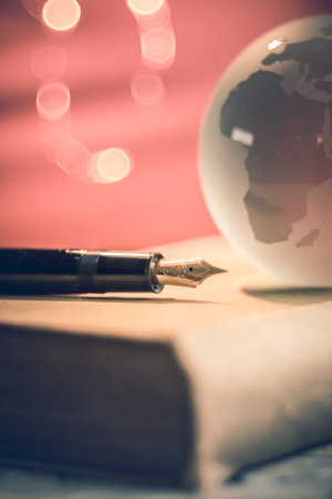 close-up of black pen and glass globe with countries on an old book. Orange background illuminated with small spot lights. writing or travel conceptの写真素材