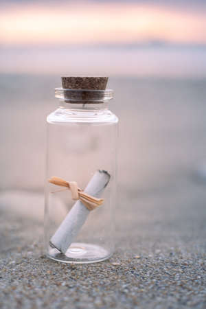 close-up view of a small glass bottle containing a paper message. the bottle is standing on the beach during sunriseの写真素材