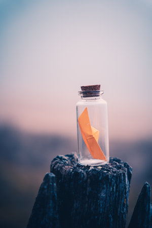 Miniature origami ship in glass bottle standing on a wooden fence at sunriseの写真素材