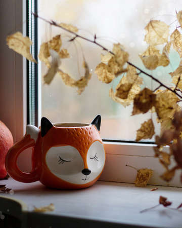 Orange mug in the shape of a fox on a windowsill with a bouquet of autumn leaves