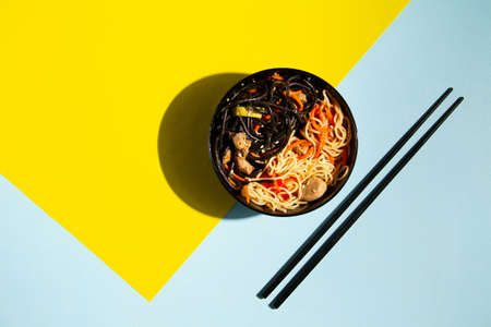 Black and white wok noodles in a black cup with black chopsticks on a bright yellow and blue background, with a hard shadow, geometry, minimalism, pin upの写真素材