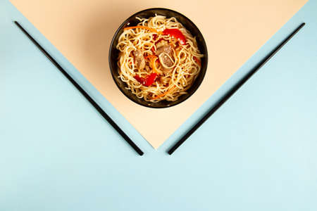 White wok noodles with meat, pepper, sesame seeds in a black plate with black chopsticks on a peach...