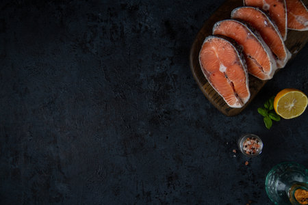 Dark textured background with free space, with the edge ingredients: salmon steaks on a board, salt, pepper, oil and lemon.の写真素材