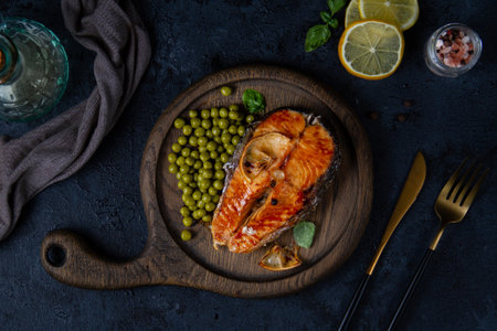 Cooked salmon steak with green peas on a wooden board for serving. Dark backdrop, a number of devices and ingredients. View from above.の写真素材