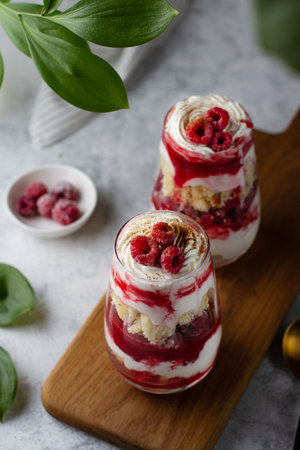 Trifle in glass glasses with raspberries and whipped cream on a table on a wooden board, green...