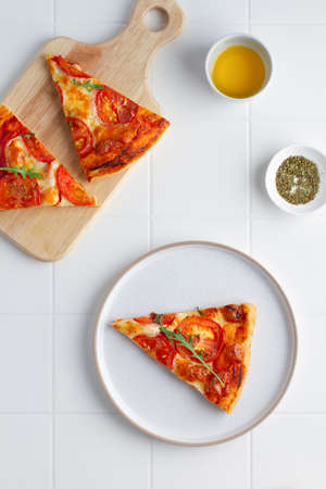 Triangular pizza slices on a plate and a board, on a white tile table, top view, minimalism