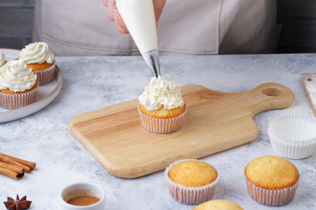 A pastry bag applies cream to a cupcake on a kitchen table with ingredients