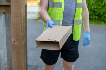 Delivery driver wearing rubber gloves handing over a parcel from a distance to avoid catching coronavirus or covid-19 during the pandemic where people are online shopping lotsの写真素材