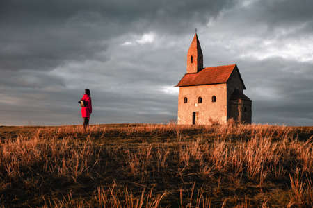 Woman tourist in red coat standing in front of Old Roman Catholic Church of St. Michael the Archangel with dark cloudy sky at Sunset - Drazovce, Slovakia.の写真素材