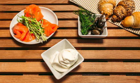 Lox, croissants, cream, capers and other ingredients prepared to cooking - top view. Traditional French croissants sandwich with fillet of salmon fish, soft cheese and capers on wooden background.の写真素材