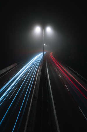 Vertical photo of cars light trails on a curved highway at night with street lamps over the road. Motion blur image -  city road with traffic headlight motion (top view) in foggy and misty weather.の写真素材