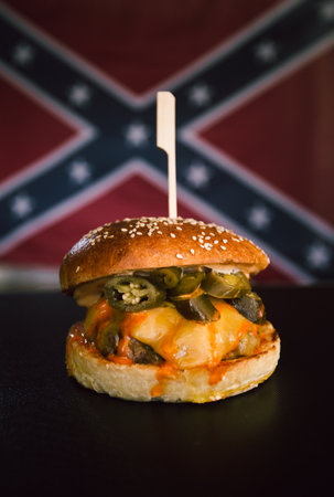 Vertical photo of american hamburger with chuck roll meat, cheddar cheeese, jalapeno etc. with confederate flag on background. DixieÂ´s vintage burgers - illuminated photo.の写真素材