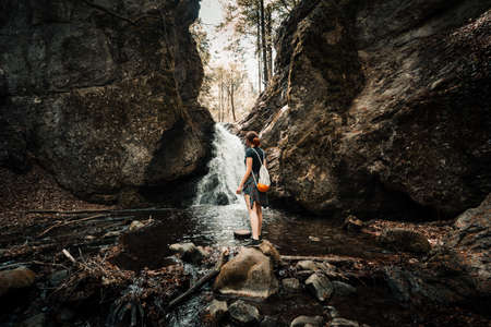 Woman (traveler and adventure) standing under the waterfall in dark and moody forest - surounded by stones and rocks. Folk tone shot of woman looking on the waterfall with backpack and relaxing.の写真素材