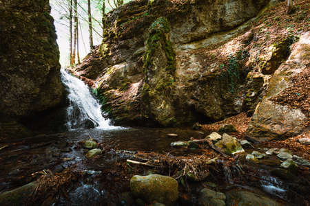 Beautiful view of moody and dramatic waterfall with big rocks and stones on foreground in forest. Small dark waterfall in European forest flows throught the tunnel of rocksの写真素材