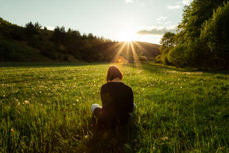 Portrait of young woman sitting in the meadow full of dandelions on sunset.  Young woman on the grass -  meadow watching and enjoying the sunset on springtime.の写真素材