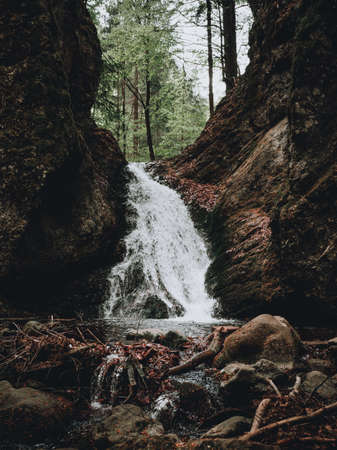Beautiful view of moody and dramatic waterfall with big rocks and stones on foreground in forest. Small dark waterfall in European forest flows throught the tunnel of rocksの写真素材