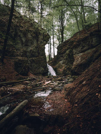 Beautiful view of moody and dramatic waterfall with big rocks and stones on foreground in forest. Small dark waterfall in European forest flows throught the tunnel of rocksの写真素材