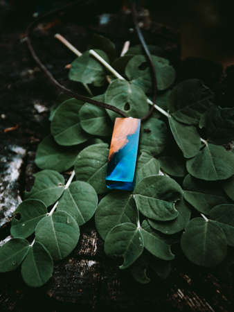 Beautiful handmaded necklace (amulet) with shining blue epoxy with green leafs on background - moody folk style photo. Handmade product of wood on wooden backround in dark forest.の写真素材