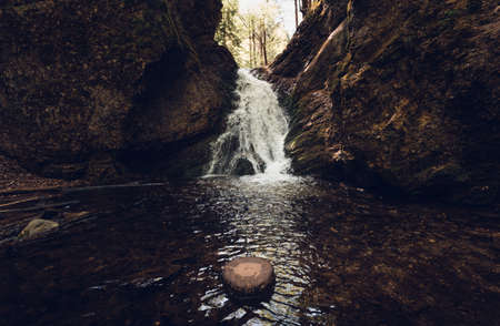 Beautiful view of moody and dramatic waterfall with big rocks and stones on foreground in forest. Small dark waterfall in European forest flows throught the tunnel of rocksの写真素材