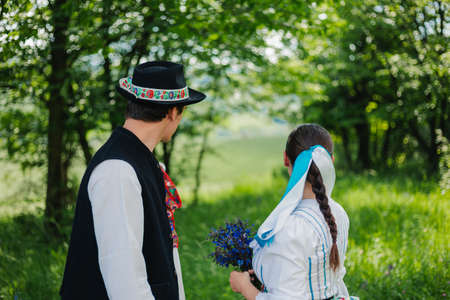 Portrait of young couple (folklore) in nature. Girl in traditional slovak and slavic dress holding a purple bouquet and looking to the forest with her lover. Couple posing to camera - horizontal shot.の写真素材