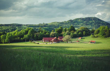 Beautiful and idyllic panorama photo of cabin (cottage or farm house) in the green meadow with forest and hills on background. Ranch in the middle of valley with cloudy sky- view from long distance.の写真素材