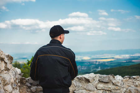 Young hiker (traveler) in black bomber jacket and cap looking and enjoying the view of landscape with city, fields and hills on background - photo from behind. Man standing on the ruins - back view.の写真素材