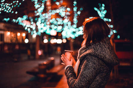 Young and beautiful woman in coat holding and drinking cup of christmas punch on christmas market in the city at night. Girl on christmas market in winter time with  a amazing lights on background.の写真素材