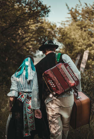 Couple of young folklorist (man and woman) walking with musical instrument and case throught the forest road in sunny day. Couple in love on forest path - folk style. Slavic Folklore  in Slovakia.の写真素材