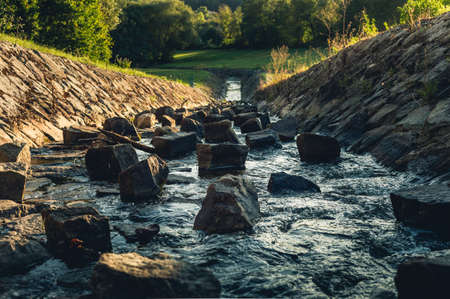 Photo of strong water stream with stones and big rocks inside flowing from reservoir (dam) in the high mountain area on sunset. Powerful current of crystal blue river with sunlight on background.の写真素材