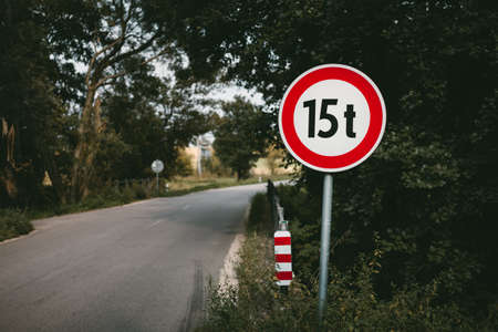 Illuminated photo of road sign (15 tons) with bridge and way throught the forest on background.  Road  (Traffic) sign of mass restriction. Red warning sign for the heavy weight vehicles with road.の写真素材