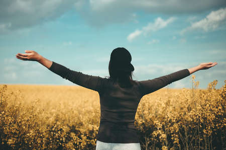 Young brunette woman spreading her hands with joy on the cole seed field with cloudy sky on background. Rear view of the woman with her arms wide spread and enjoying the sunny summer day on sunset.の写真素材