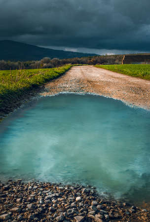 Puddle in the middle of the forest road with reflection - photo before the storm. Way in the meadow (field) and plash  with crystal blue water - mirroring effect.  Lane (path) on field on sunset.の写真素材