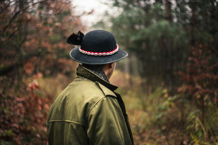 Young forester in traditional and folklore coat and hat with feather walking through the forest - back view. Photo from behind - Illuminated man with hat in green forest with foliage - dark portrait.の写真素材
