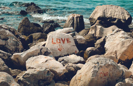 Heap of rocks and stones with writings (Love, Cioao - bye in Italian) on the beach (coast) with blue ocean on background.  Painted stone with handprint and tittle. Symbols and signs - summer Memories.の写真素材