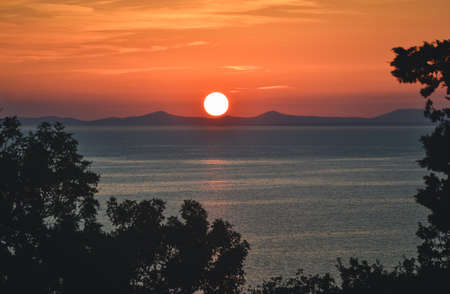 Bright sunset on the coast with big orange sun upon the hills - Image. Beautiful and amazing sunset over the sea  with yellow-orange sky and trees around the photo. View of tropical sunset on island.の写真素材