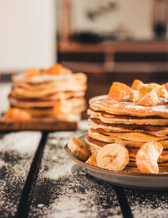Vertical photo - stack of golden pancakes with bananas and oranges on wooden board covered with caster sugar. Heap of american pancakes with maple syrup in kitchen.の写真素材