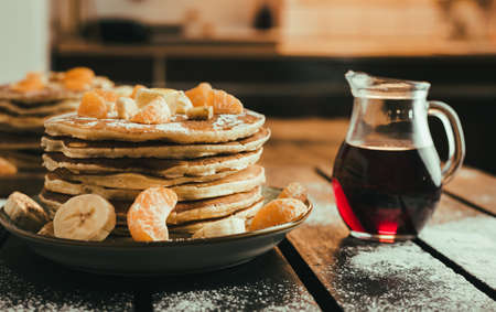 Stack of golden pancakes with bananas and oranges on wooden board covered with caster sugar. Heap of american pancakes with a jug full of syrup in kitchen.の写真素材