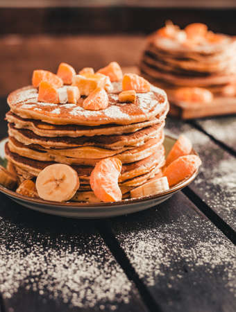 Vertical photo - stack of golden pancakes with bananas and oranges on wooden board covered with caster sugar. Heap of american pancakes with maple syrup in kitchen.の写真素材