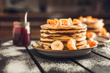 Stack of golden pancakes with bananas and oranges on wooden board covered with caster sugar. Heap of american pancakes with maple syrup and a glass of jam in kitchen.の写真素材