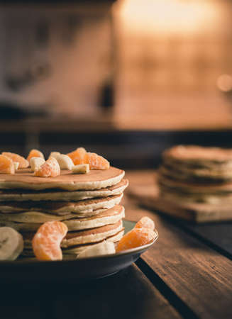Vertical photo - stack of golden pancakes with bananas and oranges on wooden board covered with caster sugar. Heap of american pancakes with maple syrup with sieve on wooden table -light on backgroundの写真素材