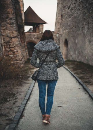 Young dark brown haired girl in a jeans and coat walking into the castle wall. Brunette woman with long legs ad long hair standing near the medieval castle with tower in autumn (view from back).の写真素材