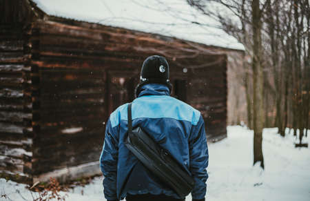 Photo of adventurer (hiker)  in snow covered forest in front of abandoned cottage (view from his back). Hiker Man in blue jacket standing in mountains with old destroyed cottage on background.の写真素材