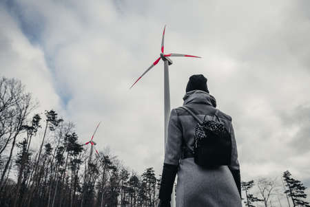 Woman standing and looking on  wind turbine station near forest  in bad weather conditions - environmental problem concept. 
Wind turbine in forest with cloudy sky - wide angle photo from below.の写真素材