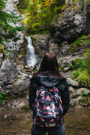Vertical photo of young and fit brunette woman with backpack looking at the waterfall with strong stream in green forest. Female tourist portrait - hiking to the mountain waterfall in autumn season.の写真素材
