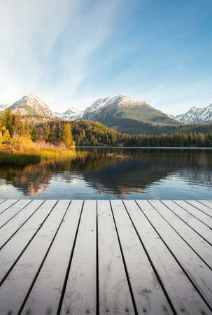 Vertical photo - pier on the lake in autumn scenery and beautiful and snowy mountains on background. Strbske pleso in High Tatras in fall with wooden mole and water reflection of landscape at sunrise.の写真素材