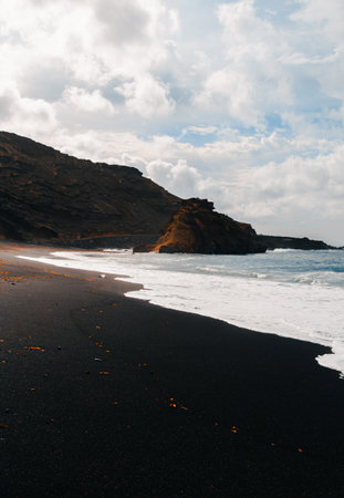 Vertical photo of black sand El Golfo beach in volcanic island Lanzarote (Canary Islands) with strong waves in blue ocean. Cliff in wild black sand beach with  cloudy blue sky on background.の写真素材