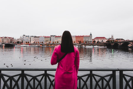 Female traveler dressed in purple coat standing near the Vltava river in Prague (Czech Republic) and looking on city - photo from her back. Wide- angle of woman tourist in city at the cold evening.の写真素材