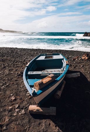 Landscape Photo of blue old boat (vessel) on volcanic black sand beach with beautiful waves in ocean on background - Lanzarote Island. Landscape shot of boat on the shore.の写真素材